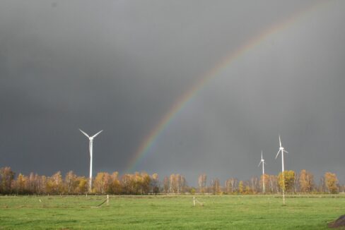 A rainbow arches over a field with several wind turbines, framed by dark storm clouds and autumn-colored trees.