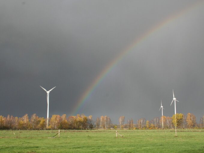 A rainbow arches over a field with several wind turbines, framed by dark storm clouds and autumn-colored trees.