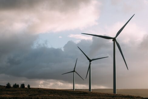 Three tall wind turbines stand on a grassy hillside under a cloudy sky, contributing to renewable energy generation.