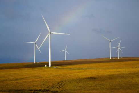 Wind turbines stand on a grassy hill under a cloudy sky, with a faint rainbow arching above them.