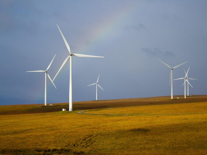 Wind turbines stand on a grassy hill under a cloudy sky, with a faint rainbow arching above them.