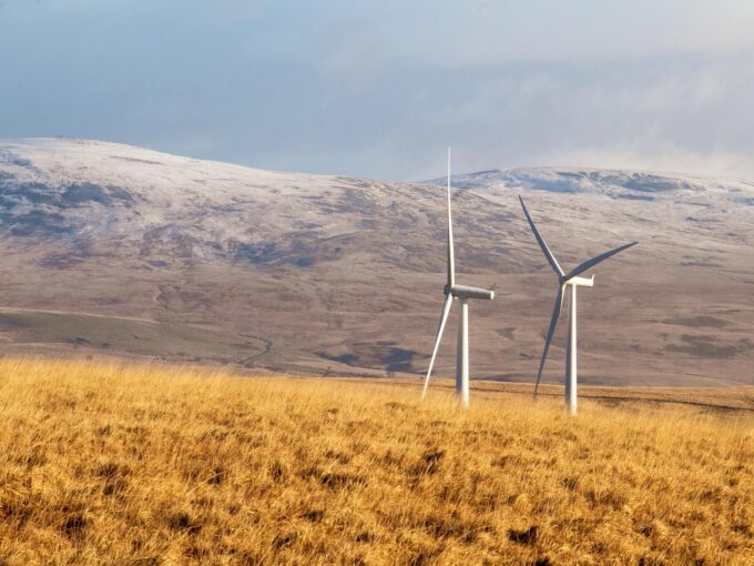 Two wind turbines stand in a golden grassy field, with snow-capped hills in the background under a cloudy sky.